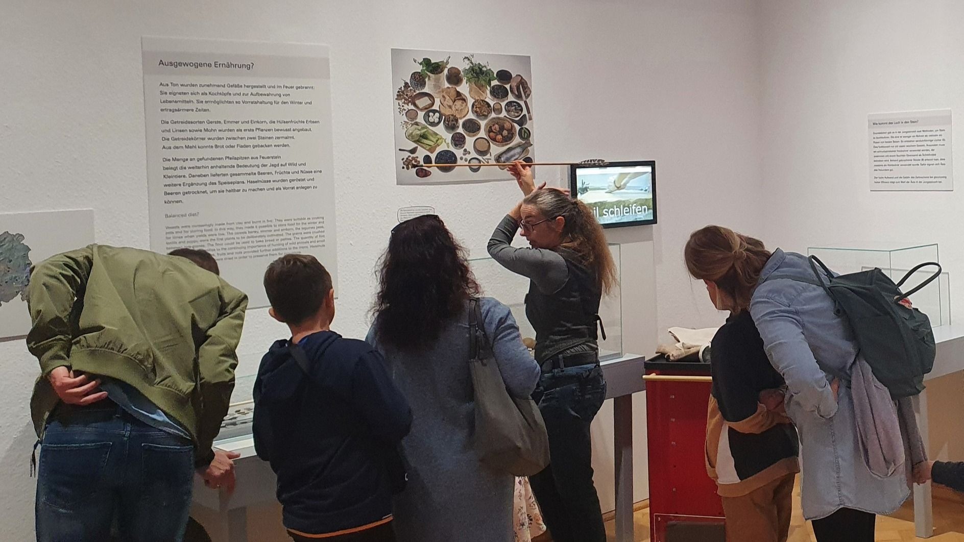 Eine Frau zeigt mit einem Speer auf eine Wandtafel mit steinzeitlichen Lebensmitteln vor einer Gruppe Besucher im Museum