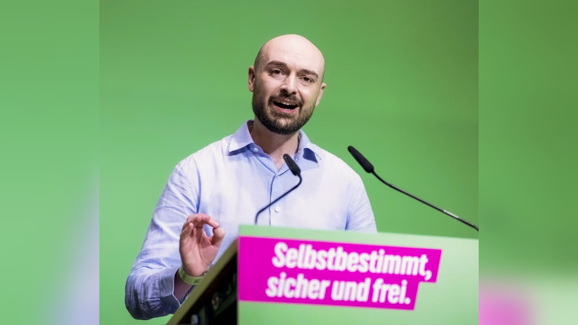 Nik Riesmeier spricht mit Bart an einem Rednerpult mit Mikrofonen vor grünem Hintergrund, Aufschrift "Selbstbestimmt,