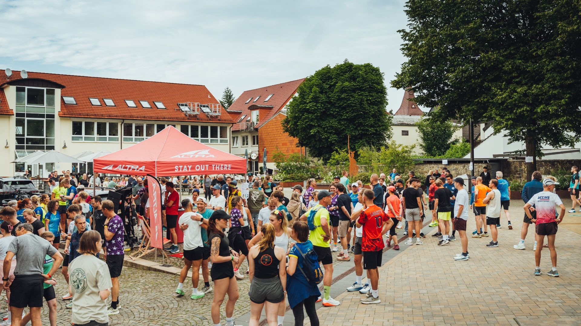 Zahlreiche Läufer und Besucher stehen auf einem Stadtplatz, umgeben von Häusern mit roten Dächern und einem roten