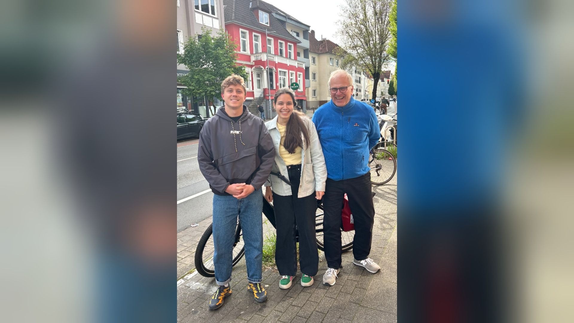 Drei lächelnde Personen, zwei junge und ein älterer Mann, stehen neben Fahrrädern auf einem Bürgersteig vor Häusern