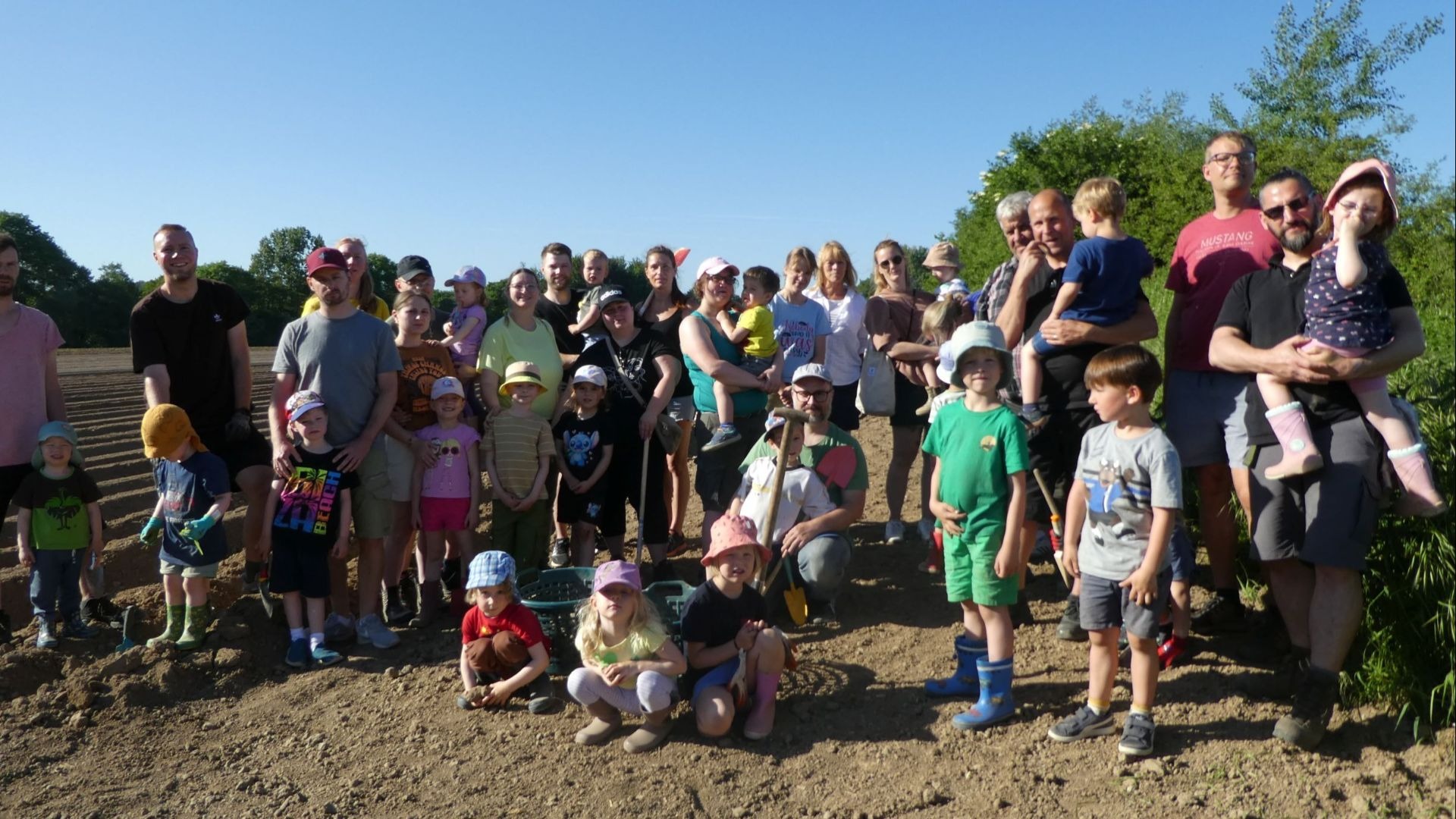 Eine große Gruppe von Erwachsenen und Kindern posiert lächelnd auf einem Kartoffelacker mit Gartengeräten unter blauem