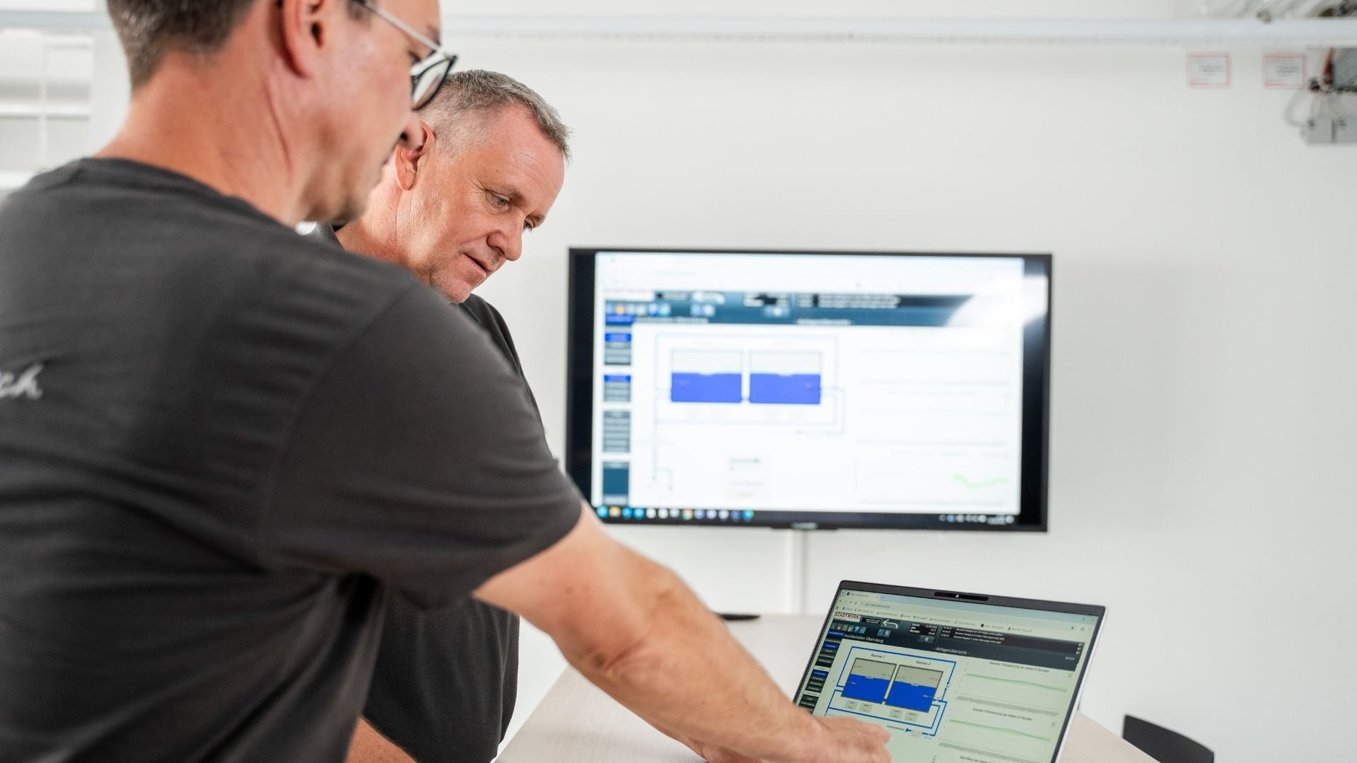 Zwei Männer schauen auf einen Laptop mit einer Grafik von Wasserstands-Anzeigen, ein Monitor zeigt ähnliche Daten