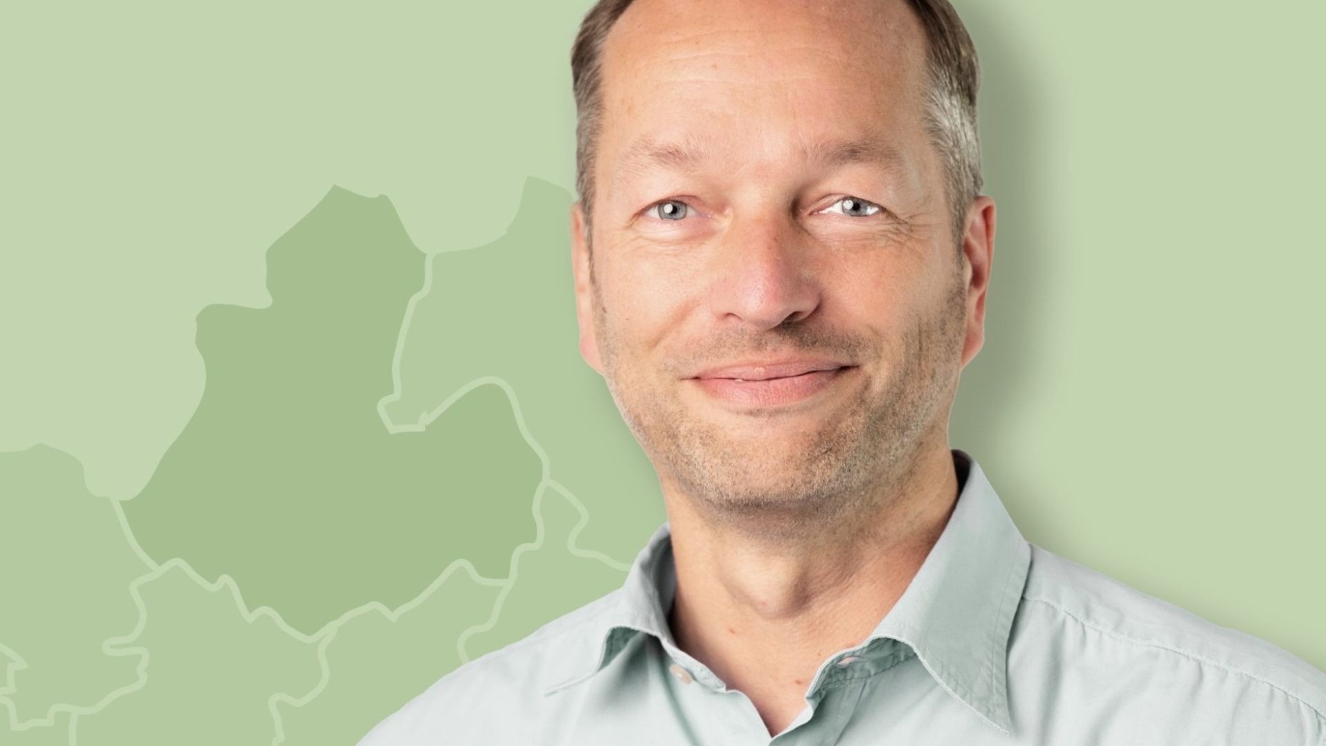 Ralf Wiebusch lächelt in einem hellblauen Hemd vor grünem Hintergrund mit schematischer Landkarte