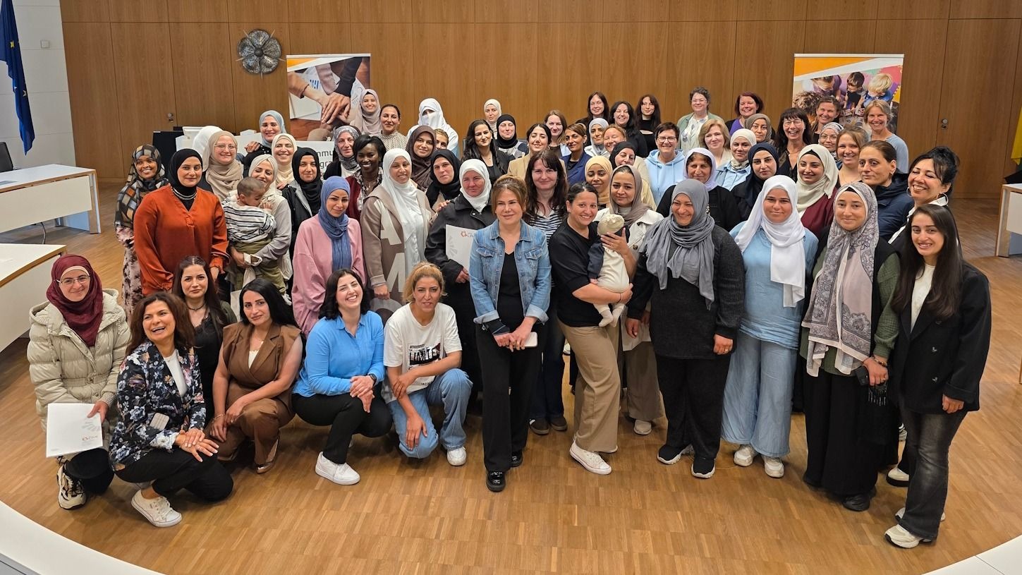 Große Gruppe lächelnder Frauen, viele mit Kopftuch, einige halten Babys, posieren in einem Raum mit Holzwänden