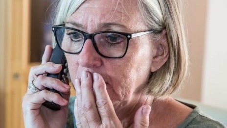 Ältere Frau mit Brille telefoniert und hält sich erschrocken die Hand vor den Mund