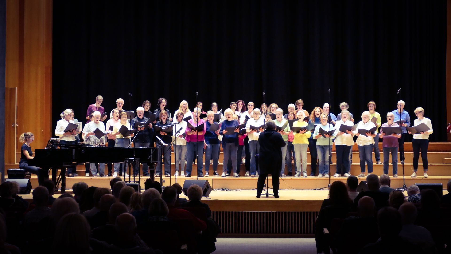 Ein großer Chor singt auf einer Bühne mit Dirigent, Pianistin und Publikum im Vordergrund in einer Konzerthalle
