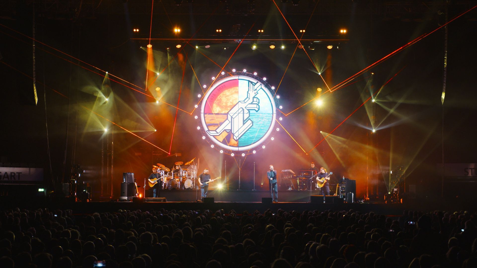 Sechs Musiker spielen auf einer großen Konzertbühne vor Publikum, hinter ihnen leuchtet ein Logo und Laserstrahlen