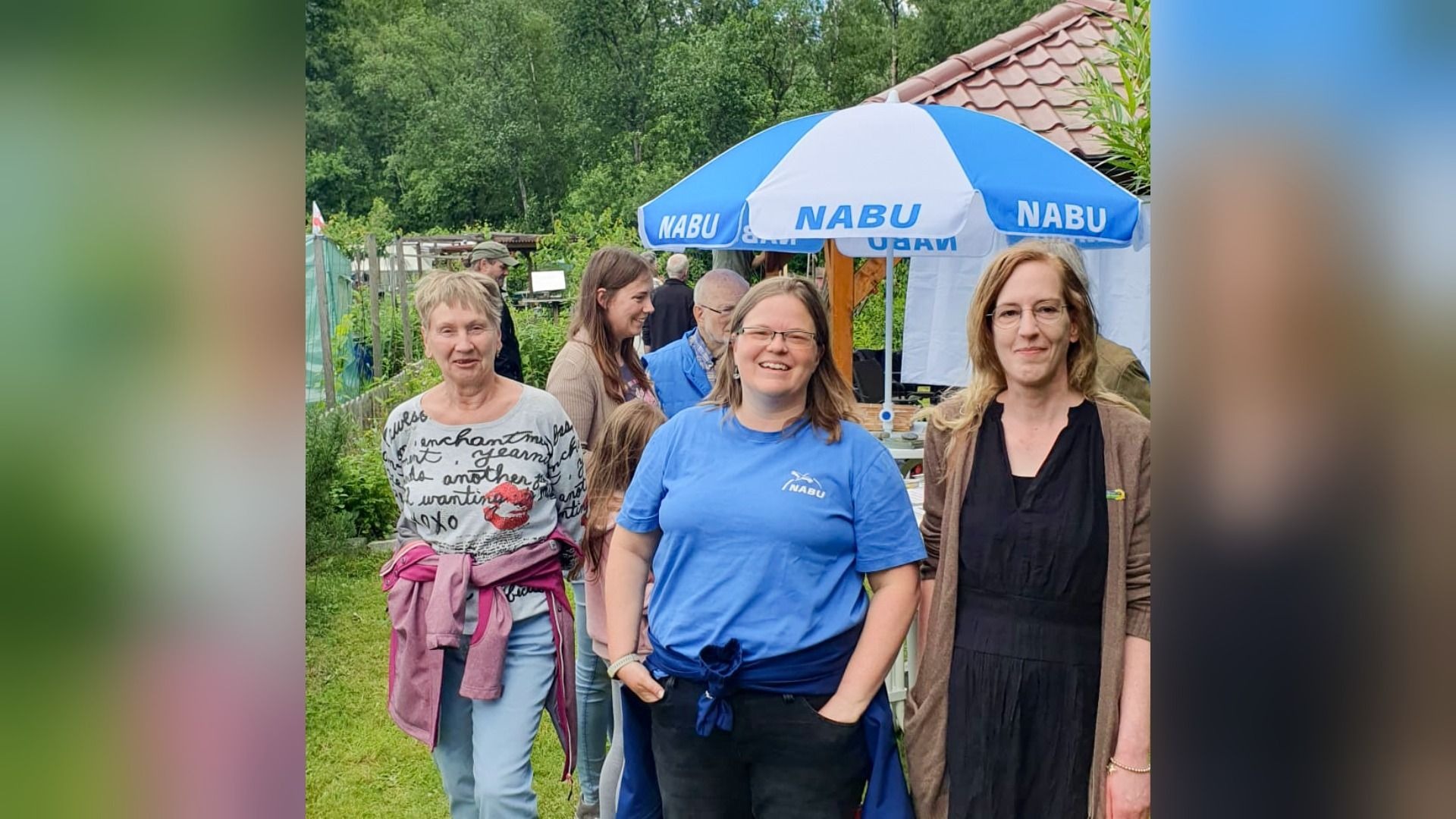Drei Frauen lächeln in die Kamera, die mittlere trägt ein blaues NABU-Shirt, dahinter ein NABU-Schirm und Menschen im