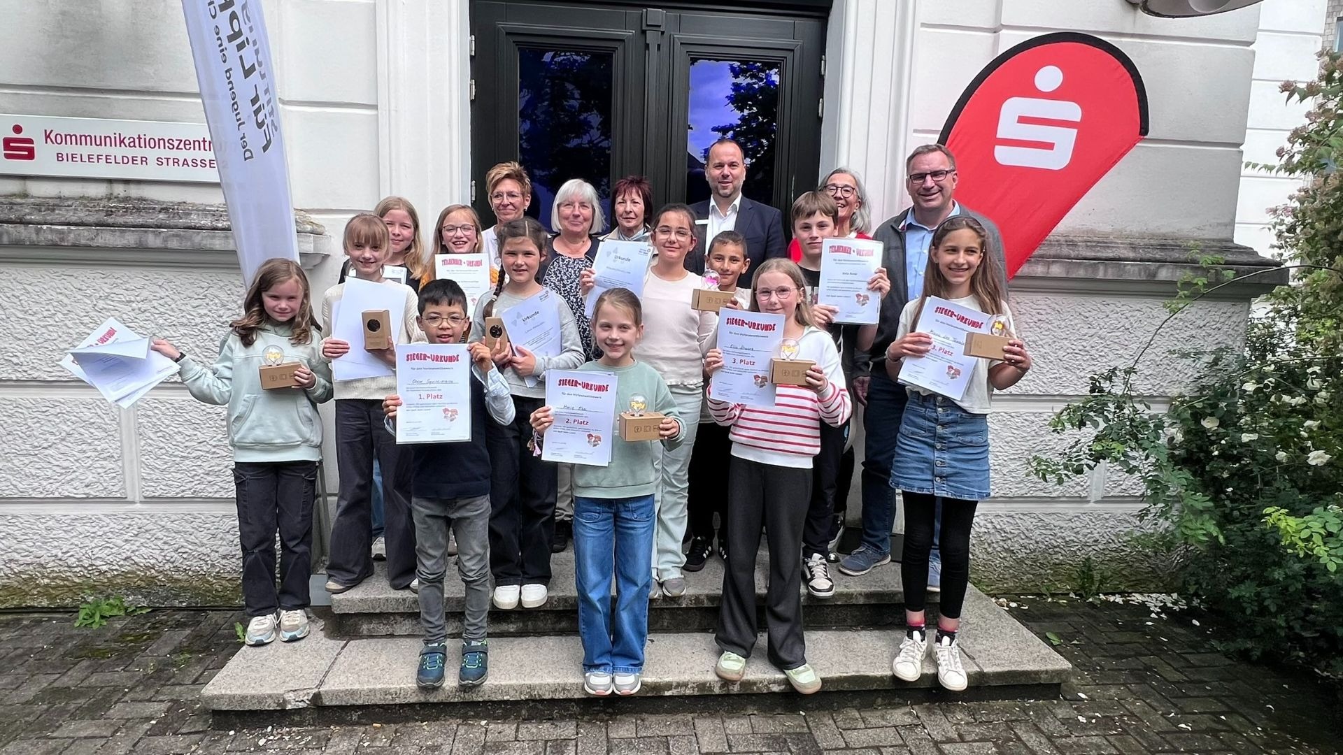 Lachende Kinder und Erwachsene posieren auf einer Treppe, Kinder halten Urkunden und Preise des Vorlesewettbewerbs