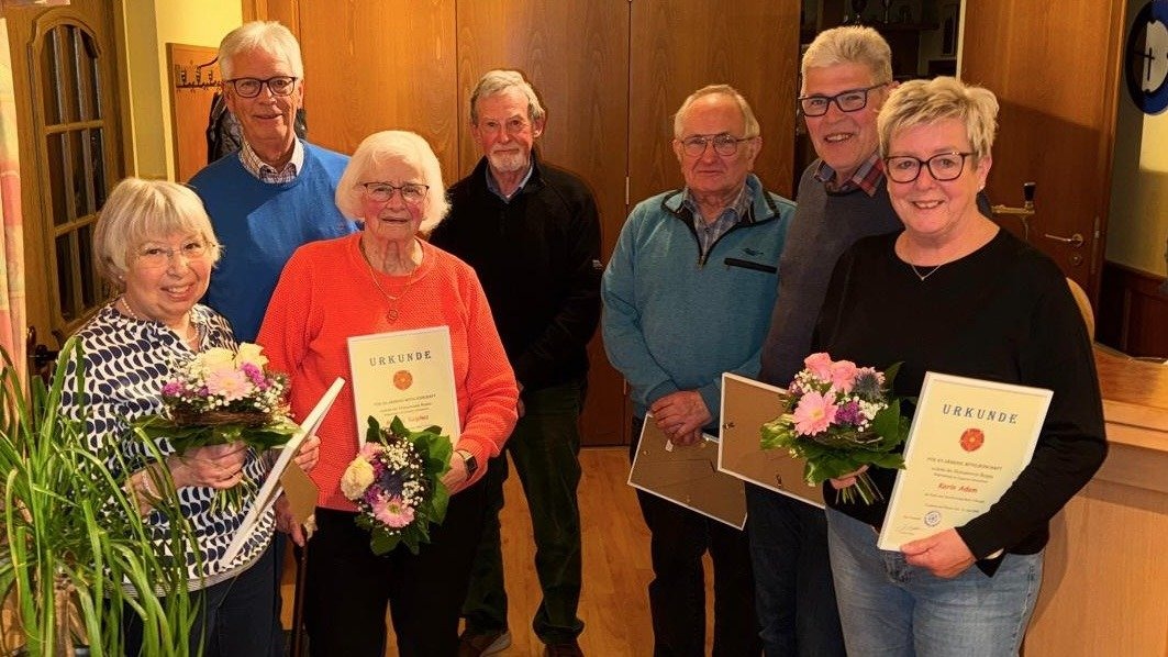 Sieben Personen, vier Männer und drei Frauen, stehen zusammen und präsentieren Urkunden und Blumensträuße