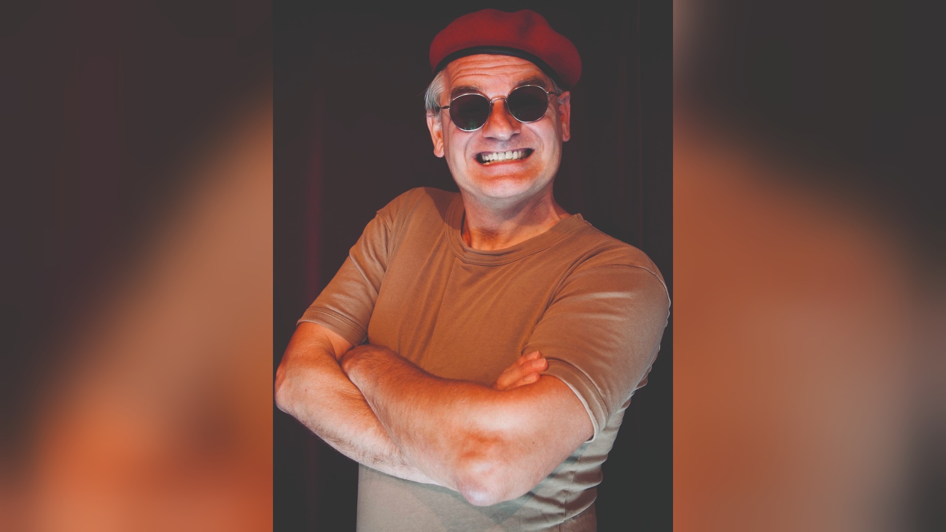 Ausbilder Schmidt mit rotem Barett, runder Sonnenbrille, beigem T-Shirt, grinsend mit verschränkten Armen