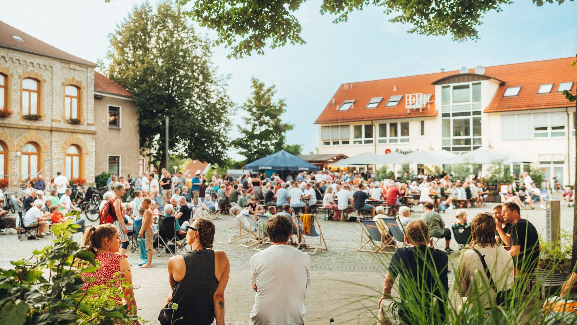 Viele Menschen sitzen an Tischen und auf Stühlen auf einem belebten Stadtplatz umgeben von Gebäuden und Bäumen