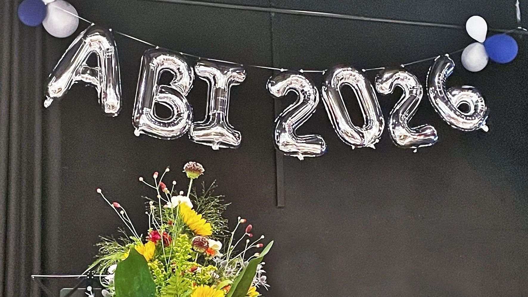 Silberne Folienballons bilden die Aufschrift "ABI 2026" über einem bunten Blumenstrauß vor dunklem Hintergrund