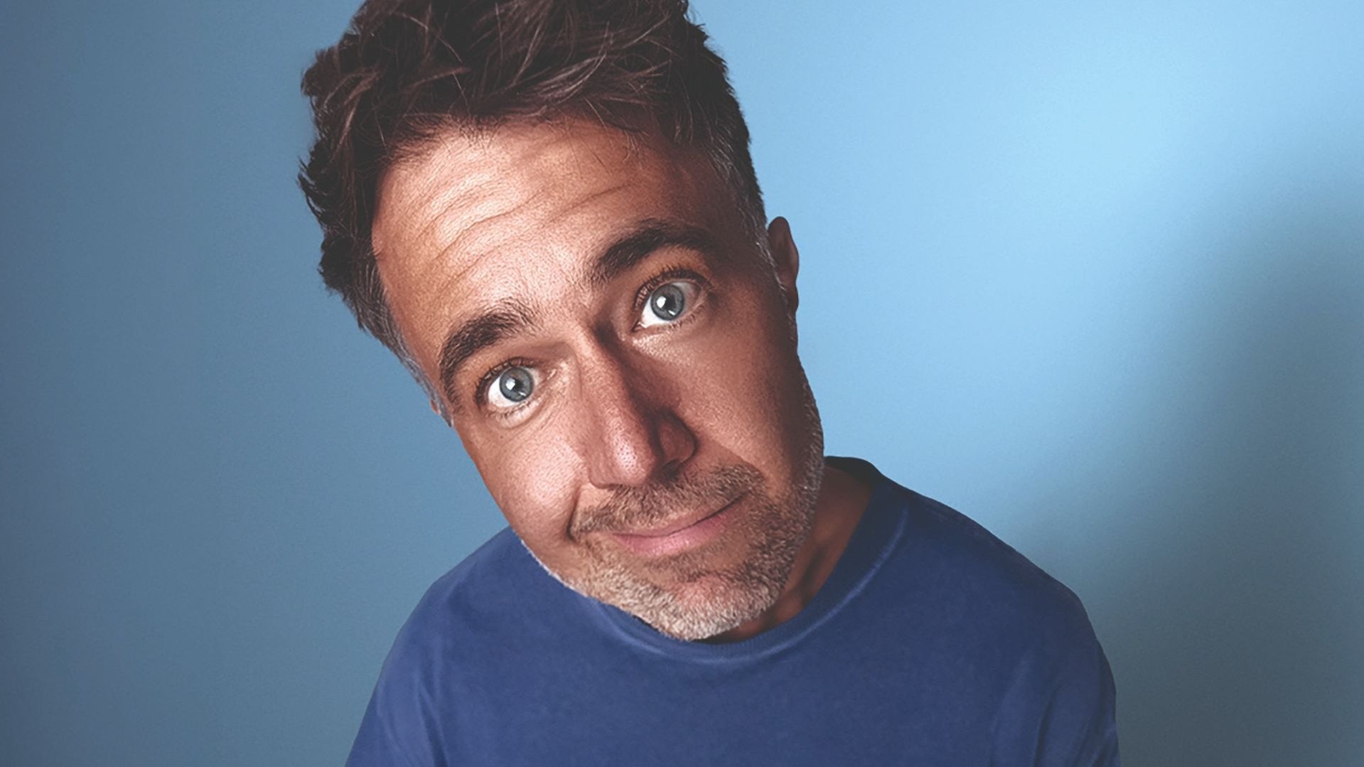 Moritz Hohl blickt mit weit geöffneten blauen Augen, leicht geneigtem Kopf und leichtem Grinsen vor blauem Hintergrund