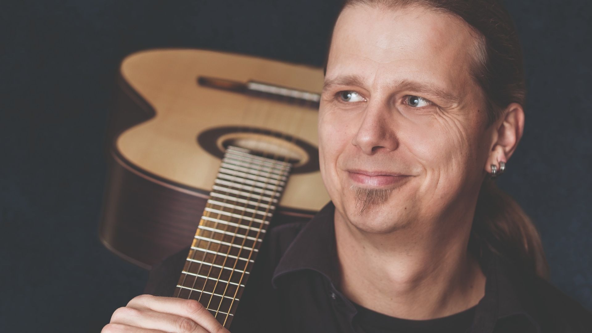 Stefan Eichner lächelt zur Seite, den Hals einer Gitarre haltend, deren Korpus im Hintergrund ist