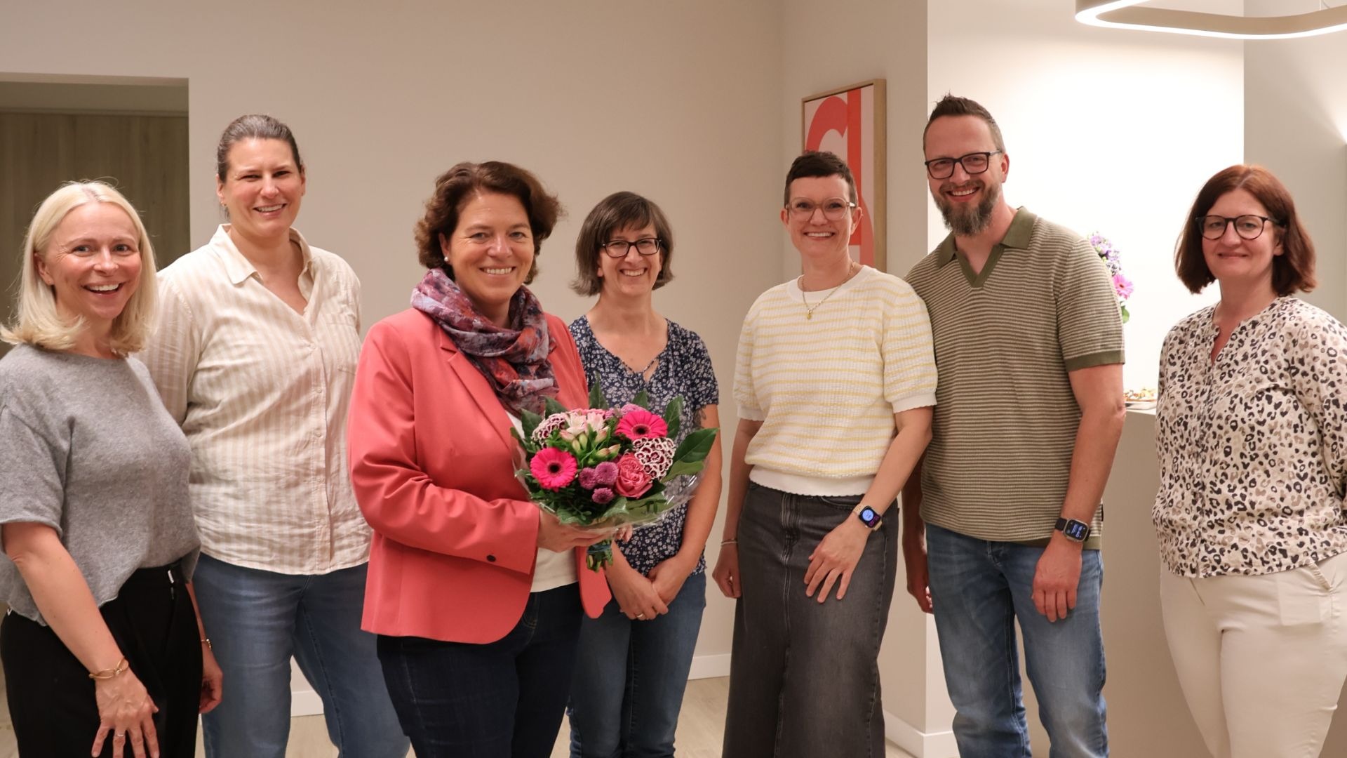 Sieben lächelnde Personen, sechs Frauen und ein Mann, stehen in einem Raum, eine Frau hält einen Blumenstrauß