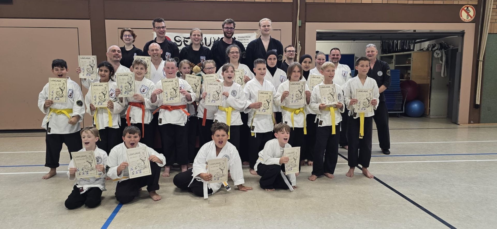 Viele Kinder in weißen Karate-Anzügen und einige Erwachsene lächeln und halten Urkunden in einer Sporthalle