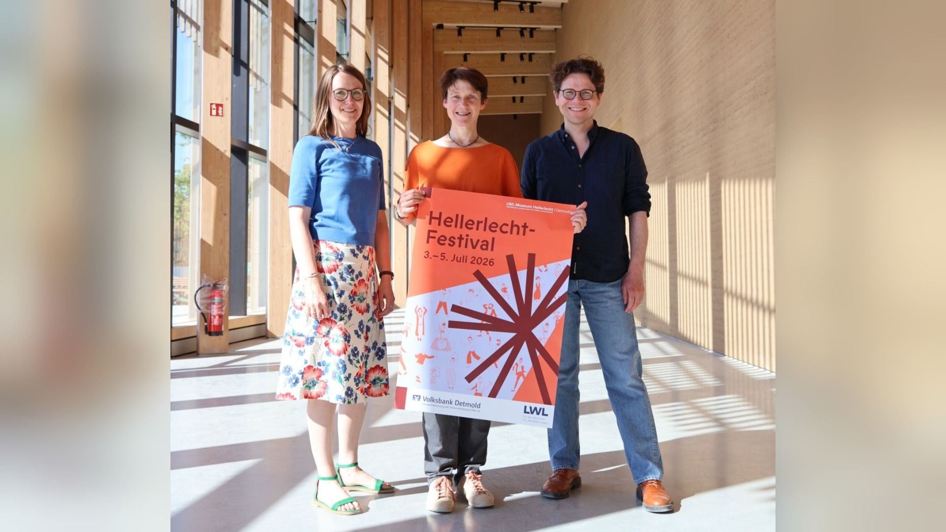 Drei Personen präsentieren ein orangefarbenes Plakat zum LWL-Hellerlecht-Festival in einem modernen Foyer