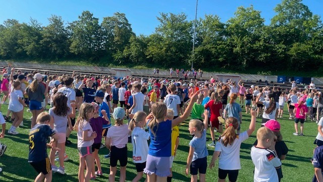Eine große Gruppe Kinder steht auf einem Sportplatz, viele haben die Arme erhoben