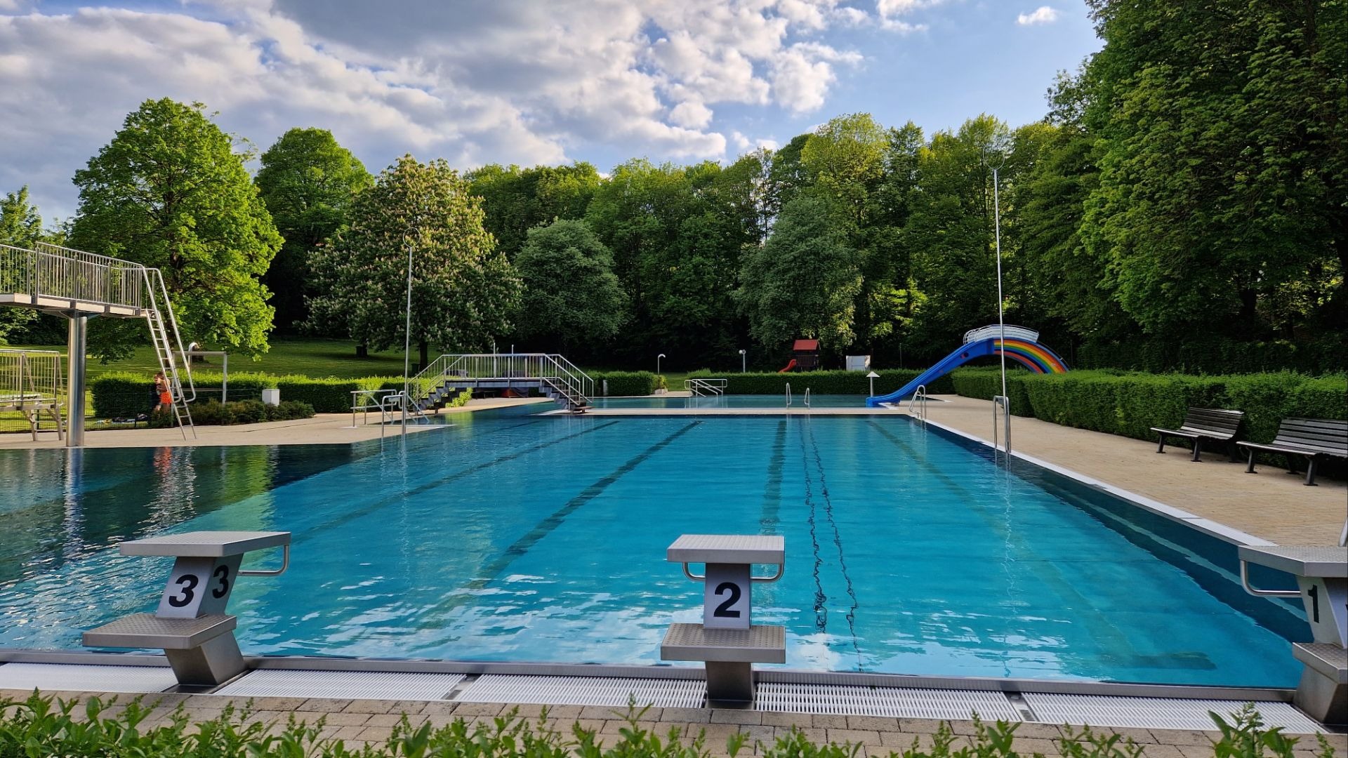 Leeres Freibad mit großem Schwimmbecken, Startblöcken, Sprunganlage, bunter Rutsche und Bäumen unter bewölktem Himmel