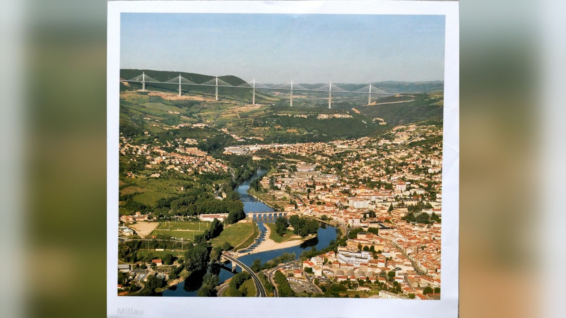 Luftaufnahme der Stadt Millau im Tal mit dem Fluss Tarn und der langen Millau-Brücke über den Hügeln