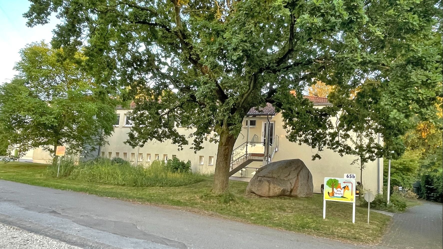 Gelbes Gebäude mit Eingangstreppe, davor großer Baum, Fels und ein Kita-Schild „Gnester 65b“ an der Straße