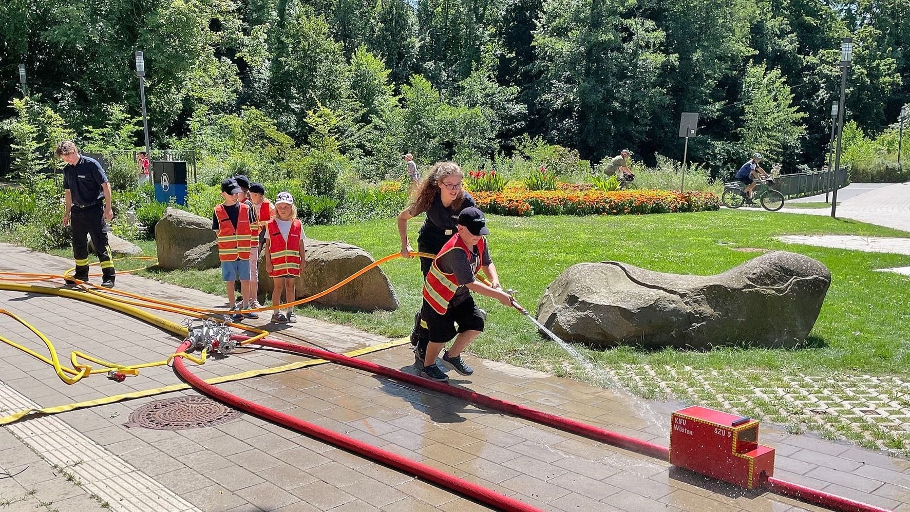 Kinderfeuerwehrübung: Ein Kind spritzt mit einem Wasserschlauch auf ein rotes Feuerwehrmodell, andere Kinder schauen zu