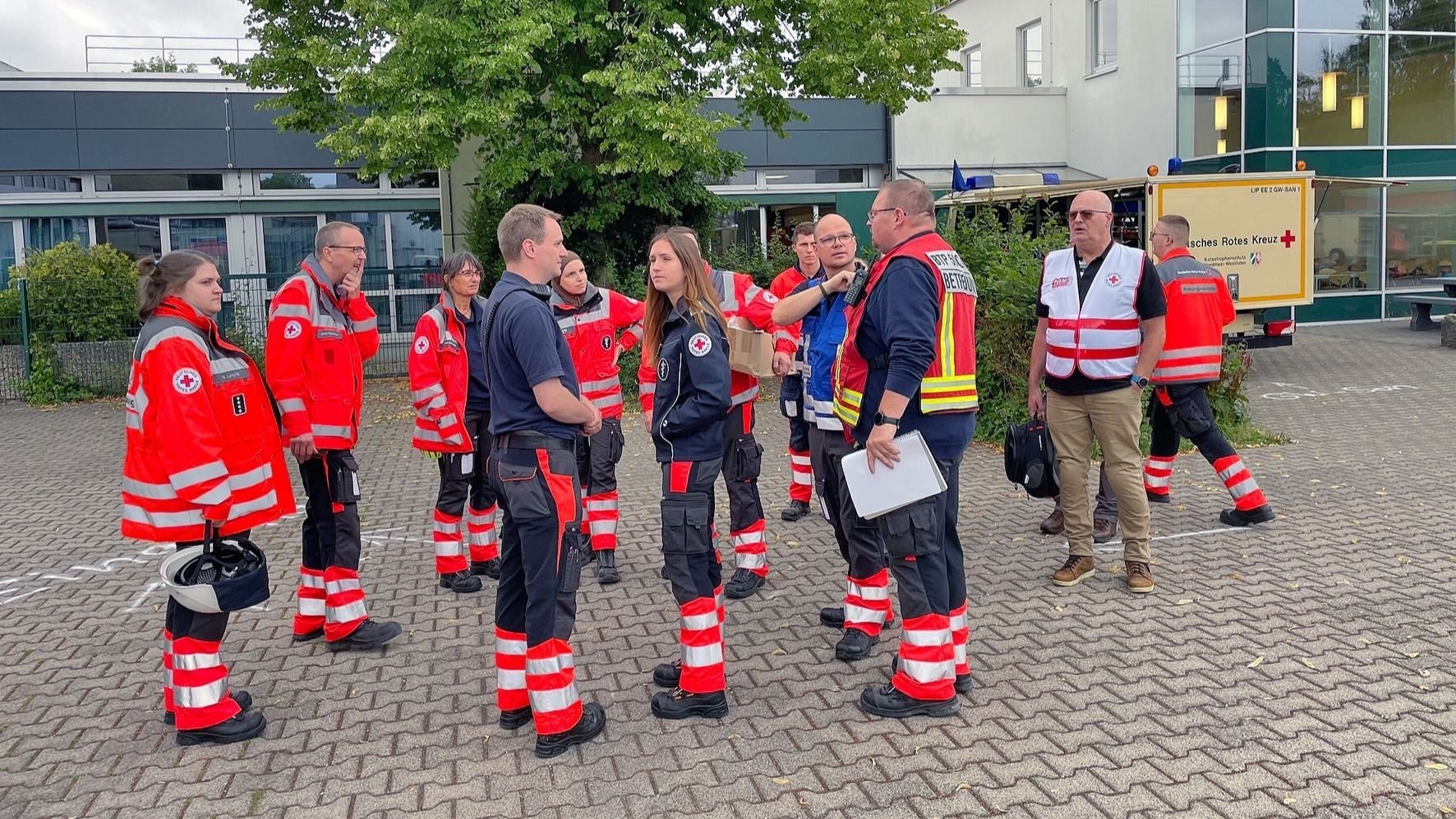 Gruppe von Einsatzkräften des Deutschen Roten Kreuzes in Uniformen spricht vor einem Gebäude und Einsatzanhänger