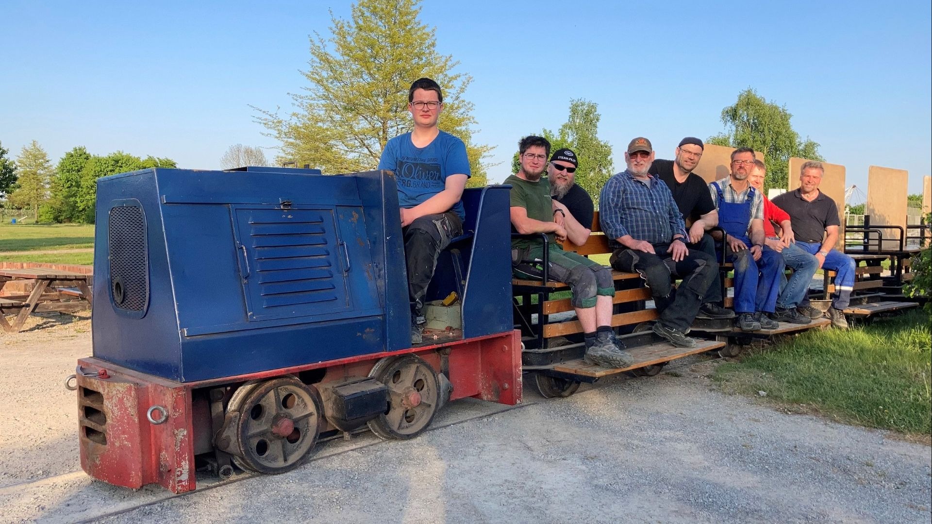 Acht Männer sitzen auf einer blauen Feldbahnlokomotive und Waggons auf Gleisen