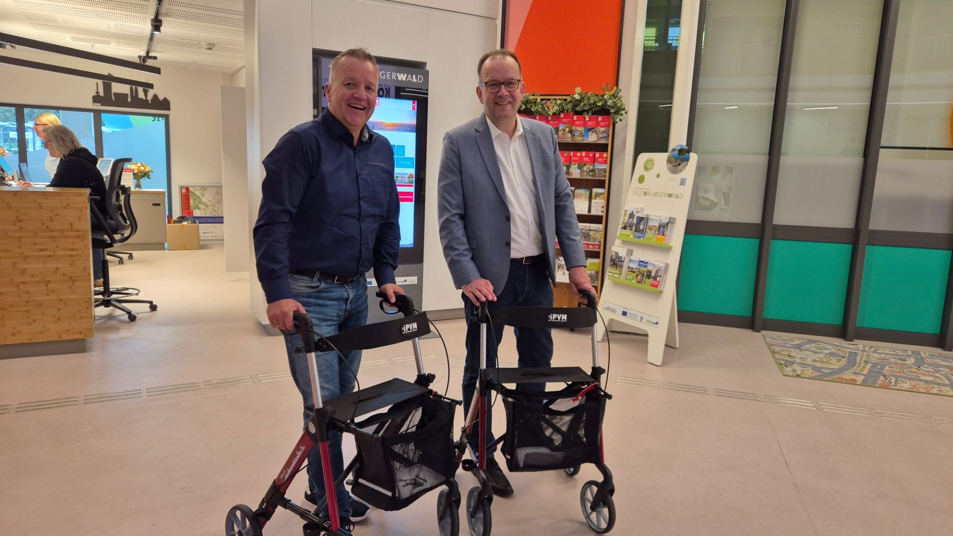 Zwei lächelnde Männer stehen in einer Tourist-Information und halten je einen Rollator