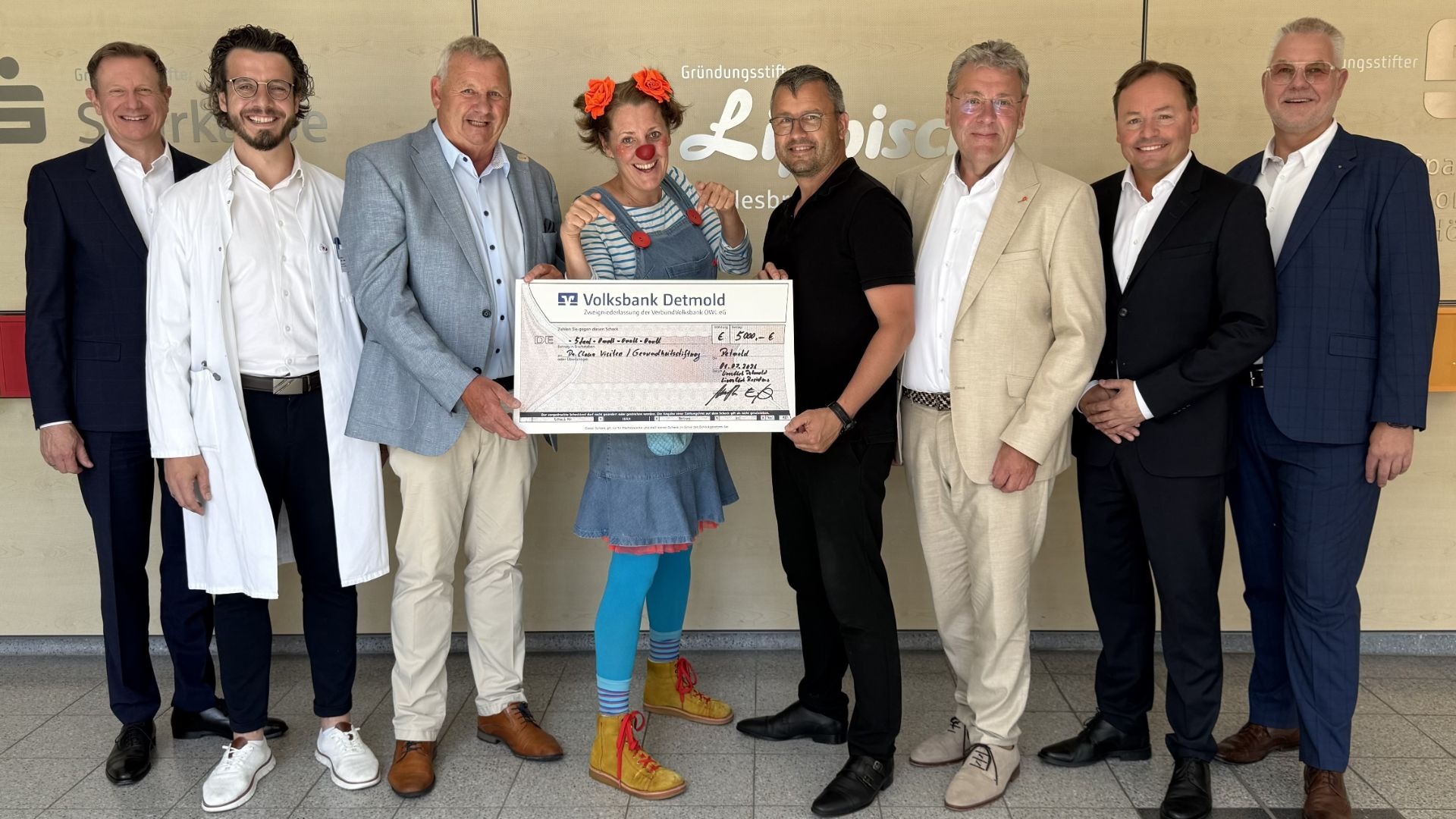 Sieben Männer und eine Clownin halten einen symbolischen Scheck für Clownsvisiten über 5000 Euro vor einer Wand mit