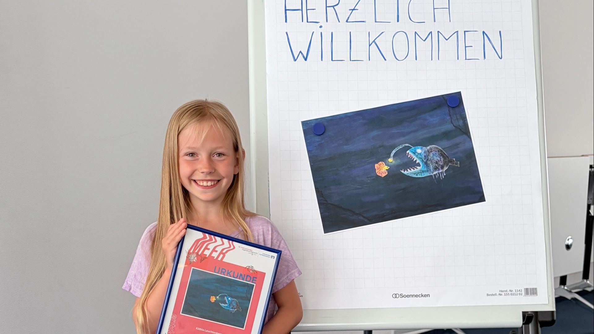 Lächelndes Mädchen hält gerahmte Urkunde mit Fischzeichnung vor Flipchart mit der Aufschrift „HERZLICH WILLKOMMEN“ und