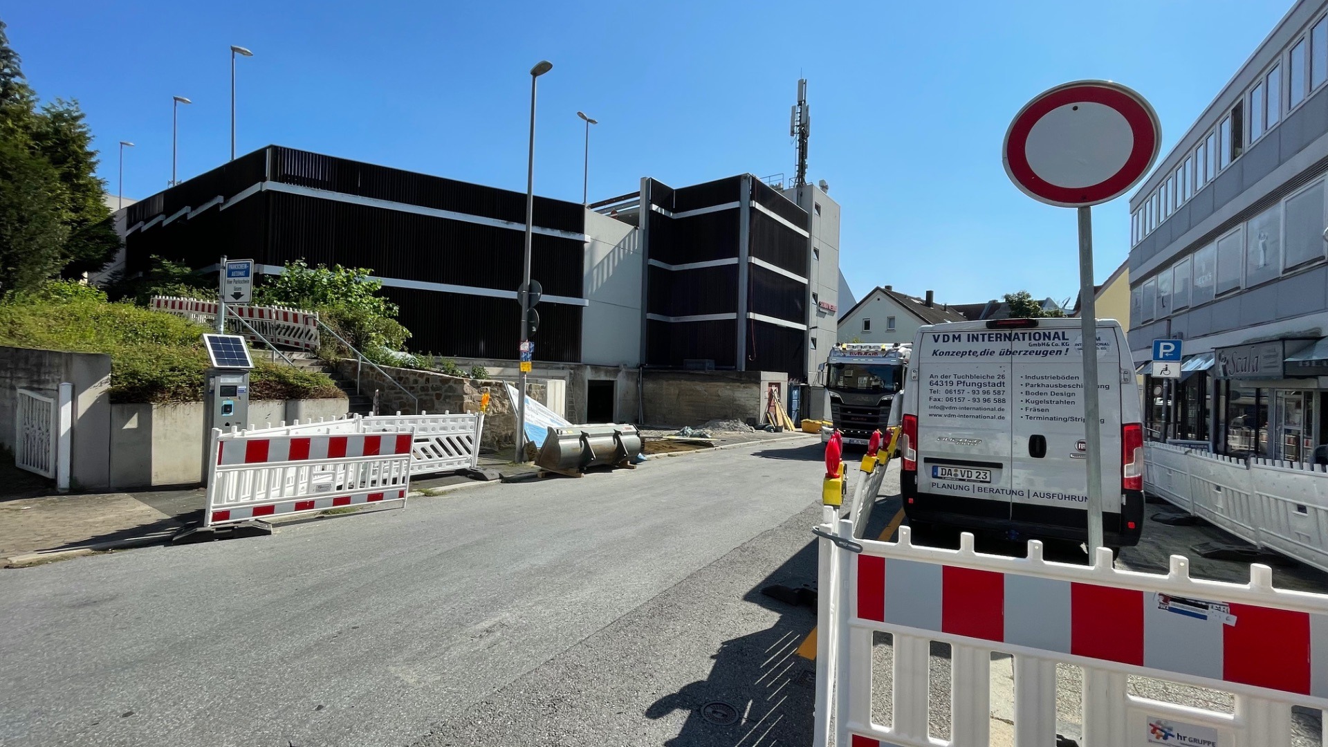 Eine Straße mit roten Bauabsperrungen, Parkscheinautomaten vor einem Parkhaus, Lieferwagen und Verbotsschild