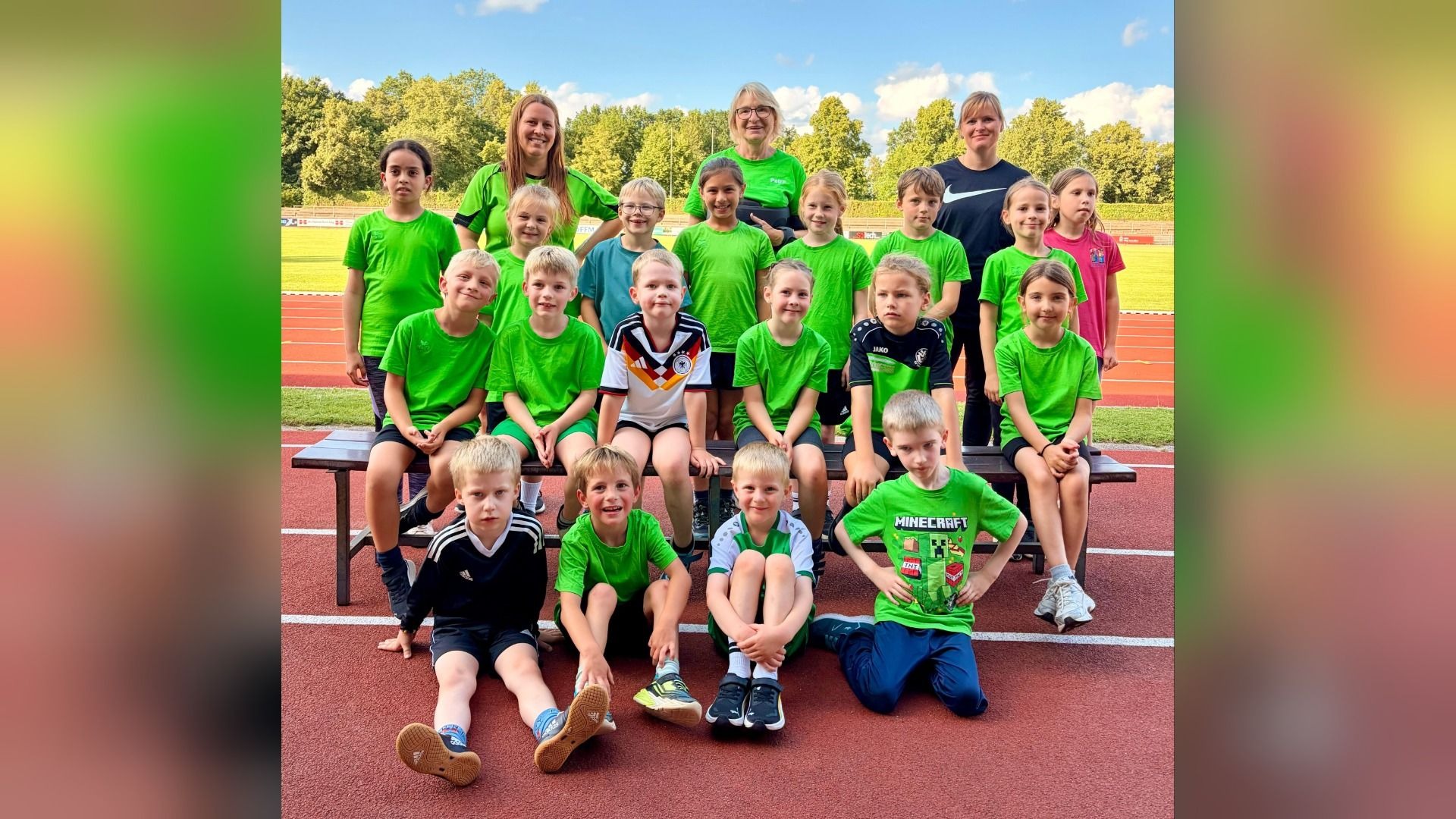 Zahlreiche Kinder und drei Frauen in grünen Sportoutfits posieren auf einer roten Laufbahn und einer Bank im Freien