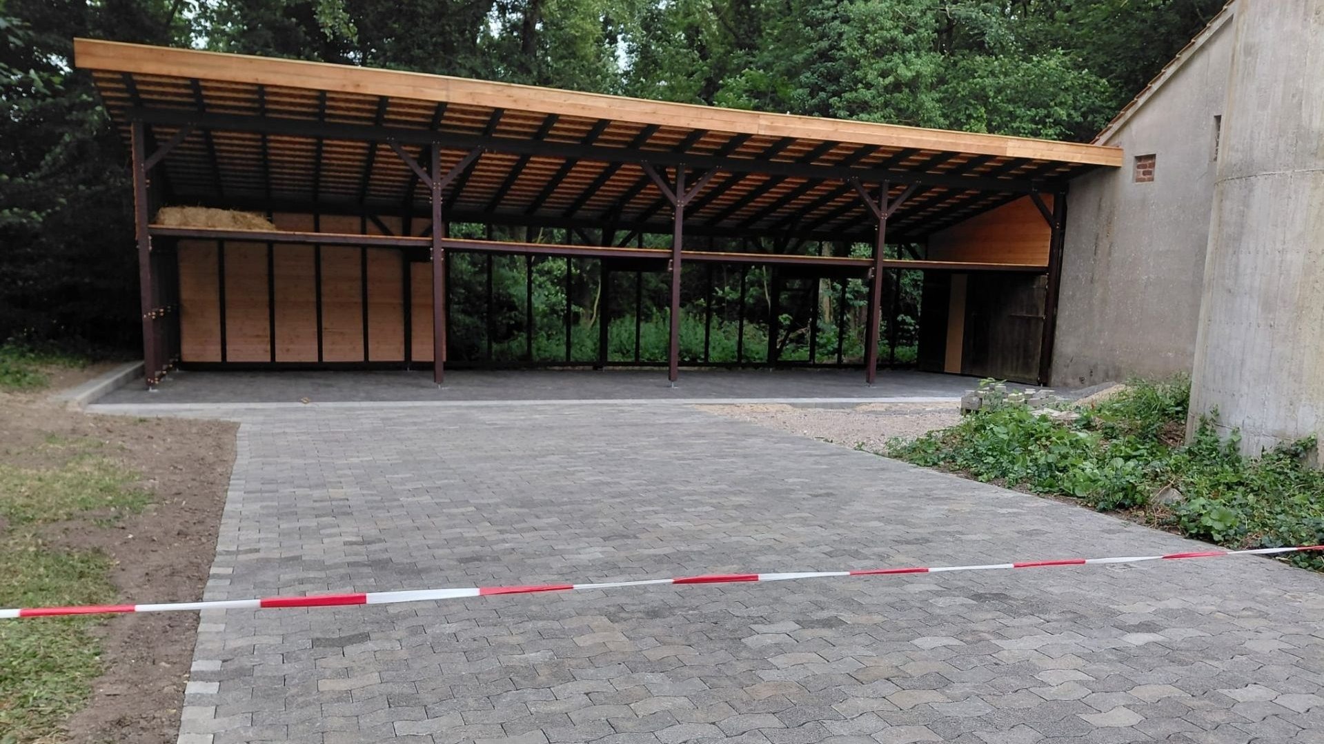 Fertiggestellte Wagenremise aus Holz mit offenem Dach und gepflastertem Vorplatz, durch rot-weißes Band gesperrt