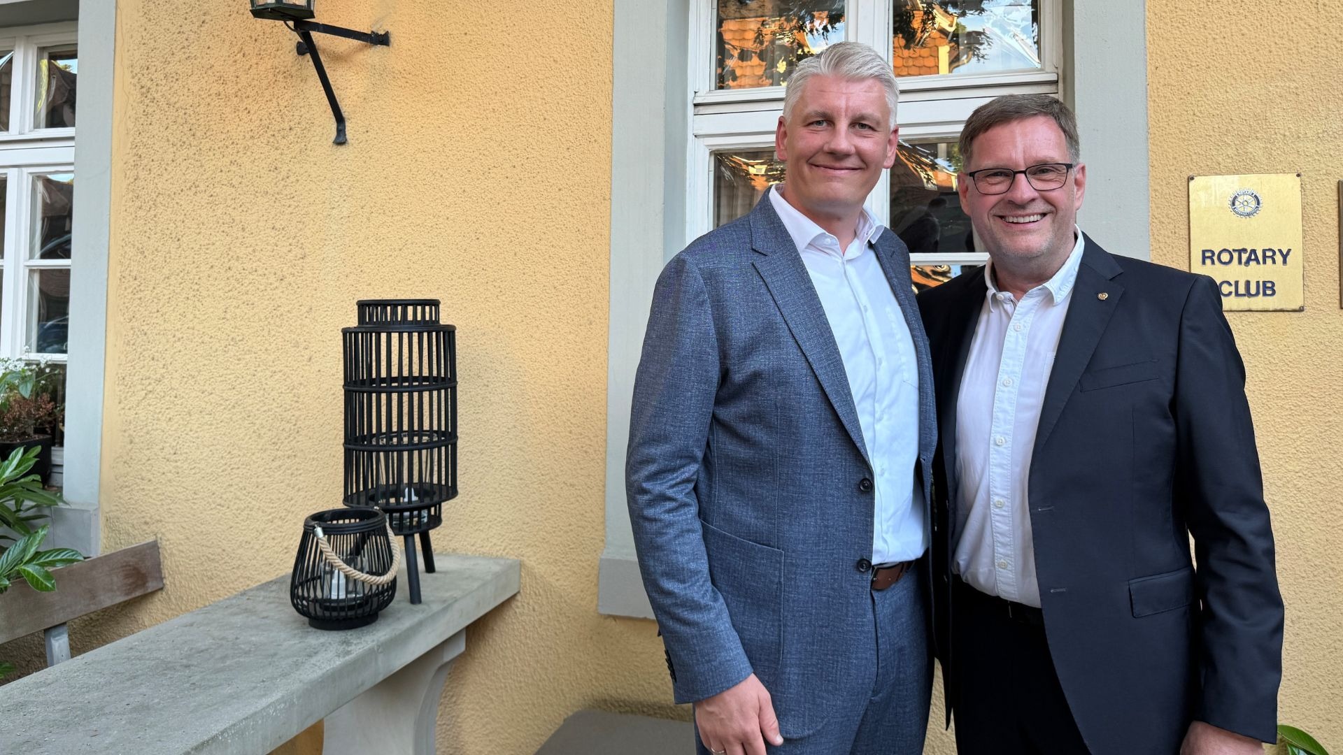Zwei lächelnde Männer in dunklen Anzügen, einer mit Brille, stehen vor gelber Hauswand und Rotary Club Schild