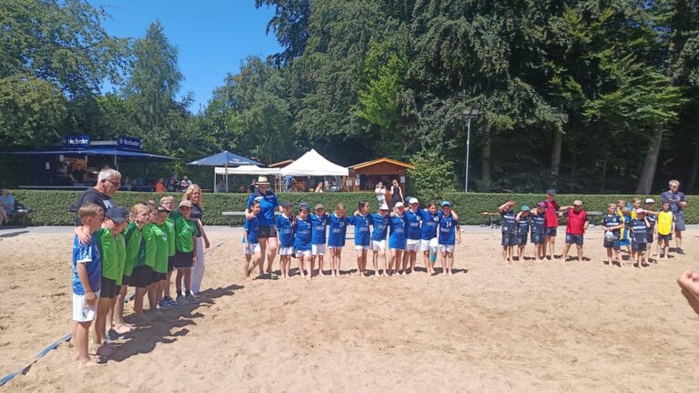 Mehrere Kindergruppen in Sporttrikots stehen mit Erwachsenen auf einem Sandfeld beim Beachhandball