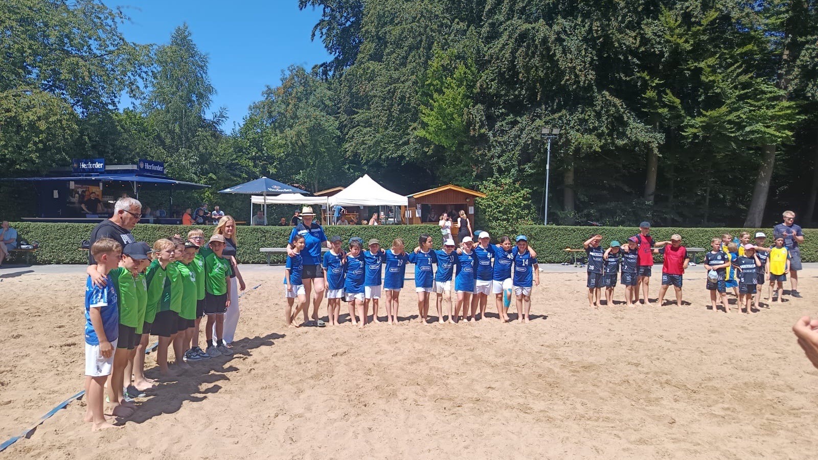 Mehrere Kindergruppen in Sporttrikots stehen mit Erwachsenen auf einem Sandfeld beim Beachhandball