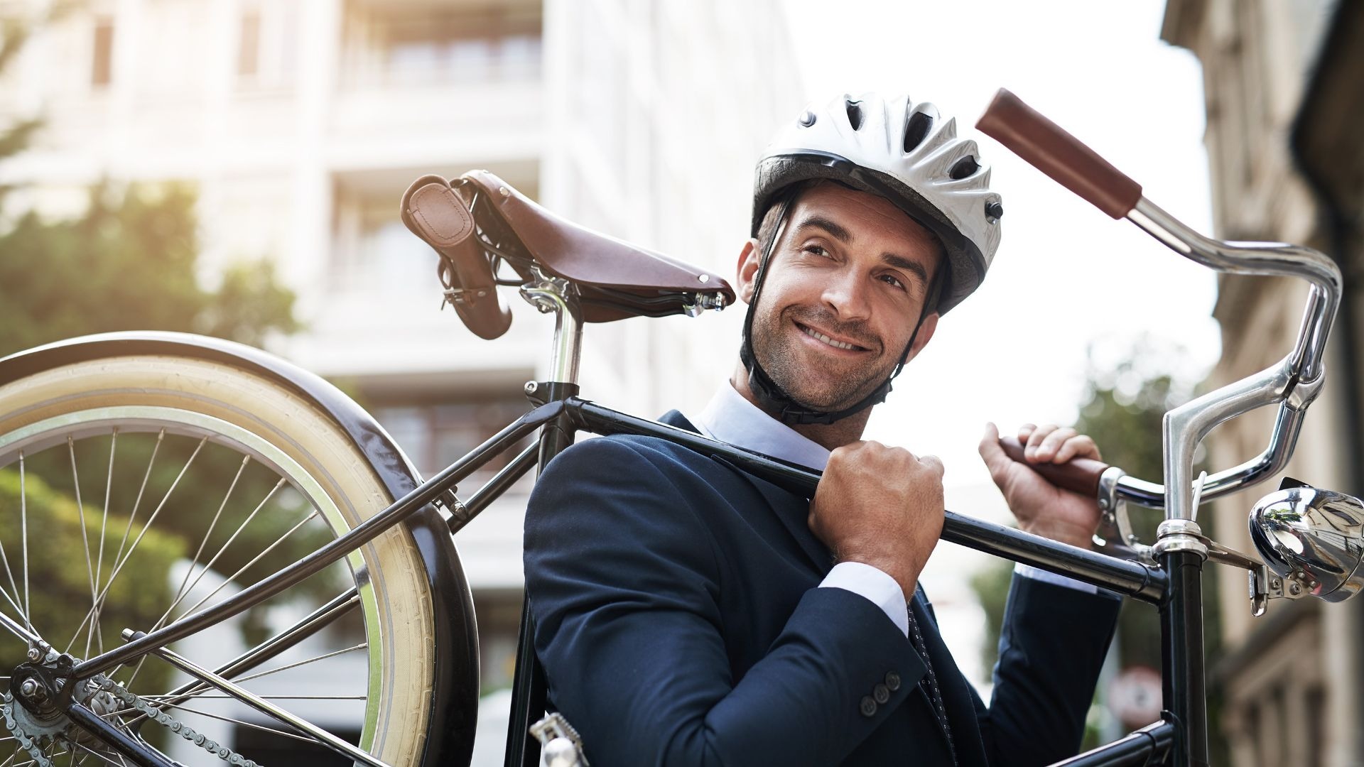 Ein lächelnder Mann mit Anzug und Fahrradhelm trägt ein Fahrrad auf der Schulter in der Stadt