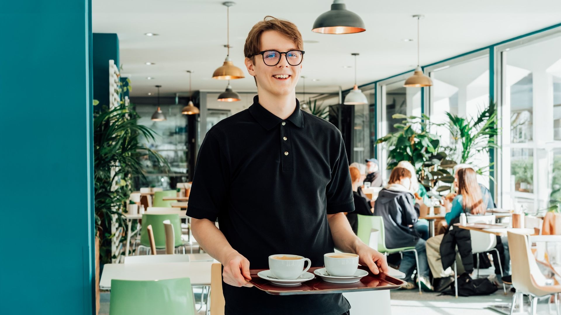 Lächelnder junger Mann mit Brille serviert zwei Kaffeetassen auf einem Tablett in einem Café