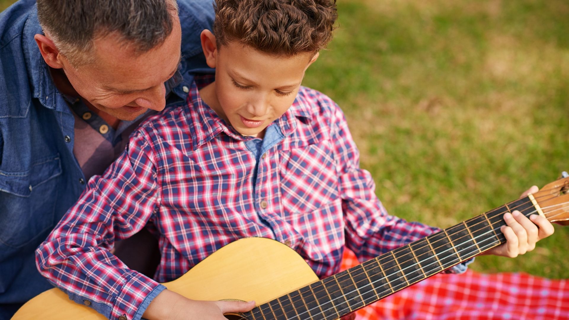 Junge spielt Gitarre, während ein Mann daneben auf einer karierten Decke im Gras sitzt