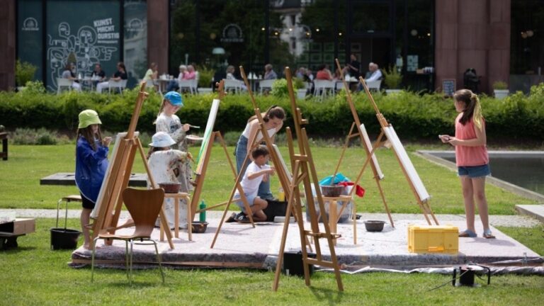 Fünf Kinder und eine erwachsene Frau malen an Staffeleien im Park vor einem Gebäude, weitere Personen im Hintergrund