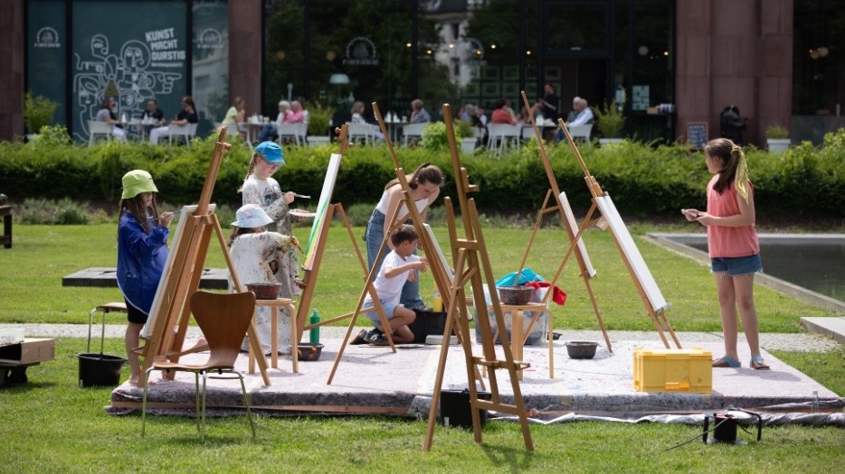 Fünf Kinder und eine erwachsene Frau malen an Staffeleien im Park vor einem Gebäude, weitere Personen im Hintergrund