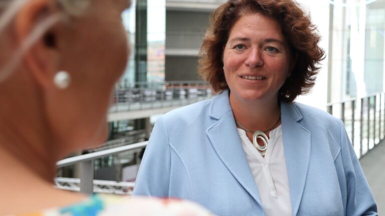 Kerstin Vieregge mit braunem Haar und blauem Blazer lächelt im Gespräch vor moderner Architektur