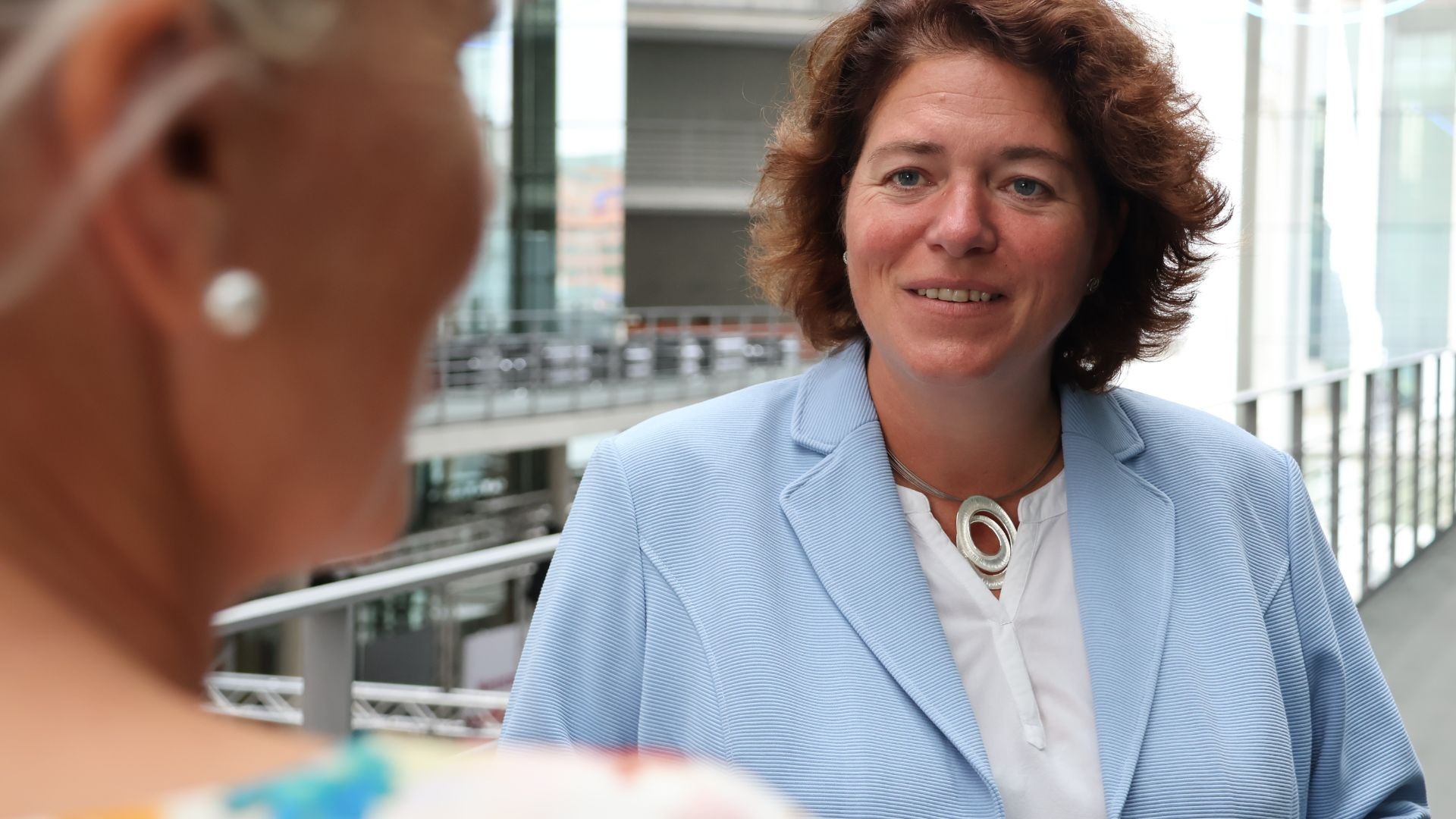 Kerstin Vieregge mit braunem Haar und blauem Blazer lächelt im Gespräch vor moderner Architektur
