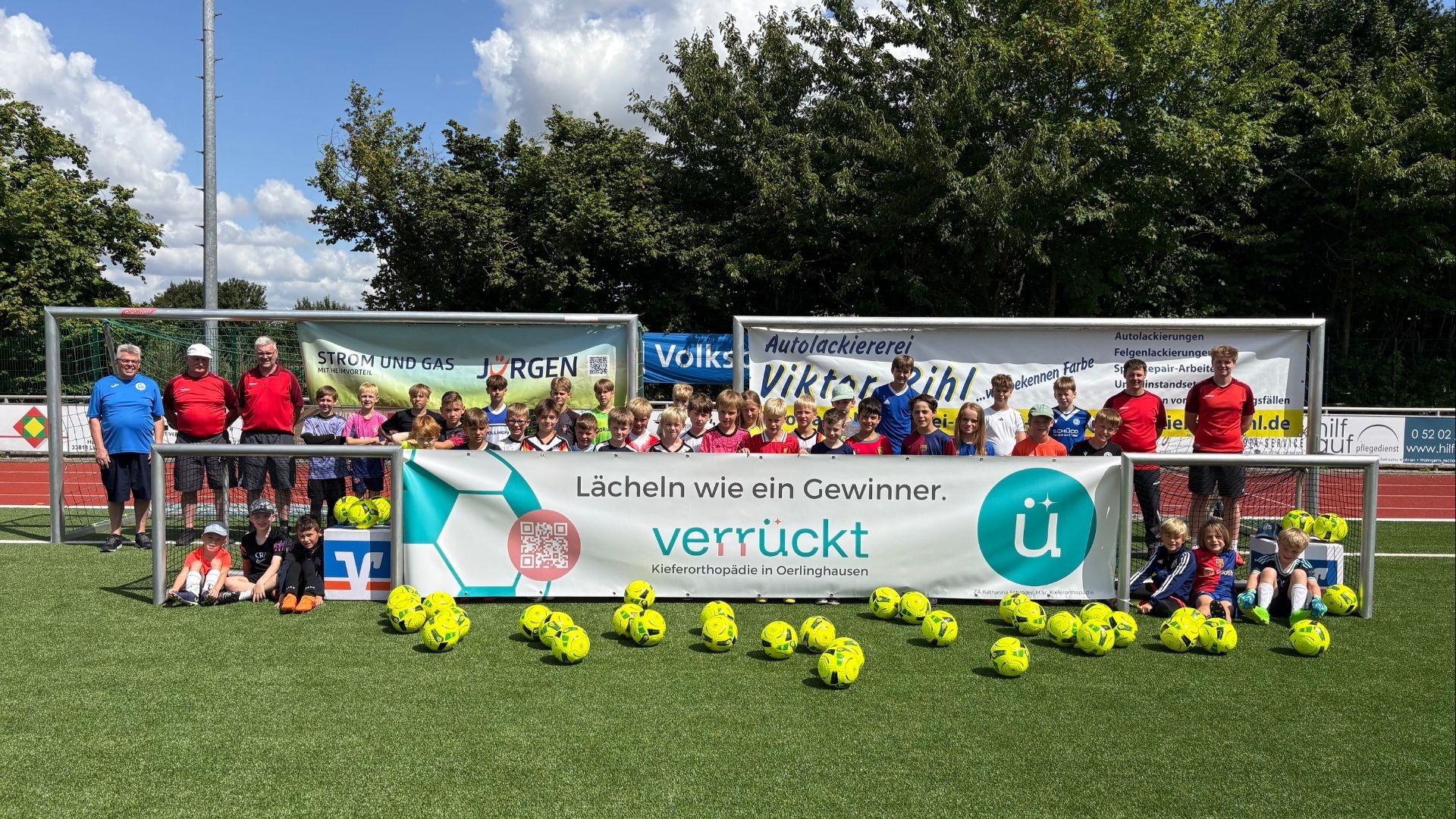 Kinder und Betreuer eines Fußballcamps posieren auf einem Fußballplatz mit vielen gelben Bällen und Sponsorbannern