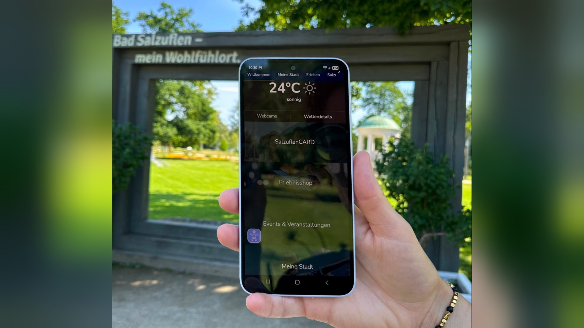 Hand hält Smartphone mit Bad Salzuflen App, die Wetter und SalzuflenCARD anzeigt, vor einem Parkschild