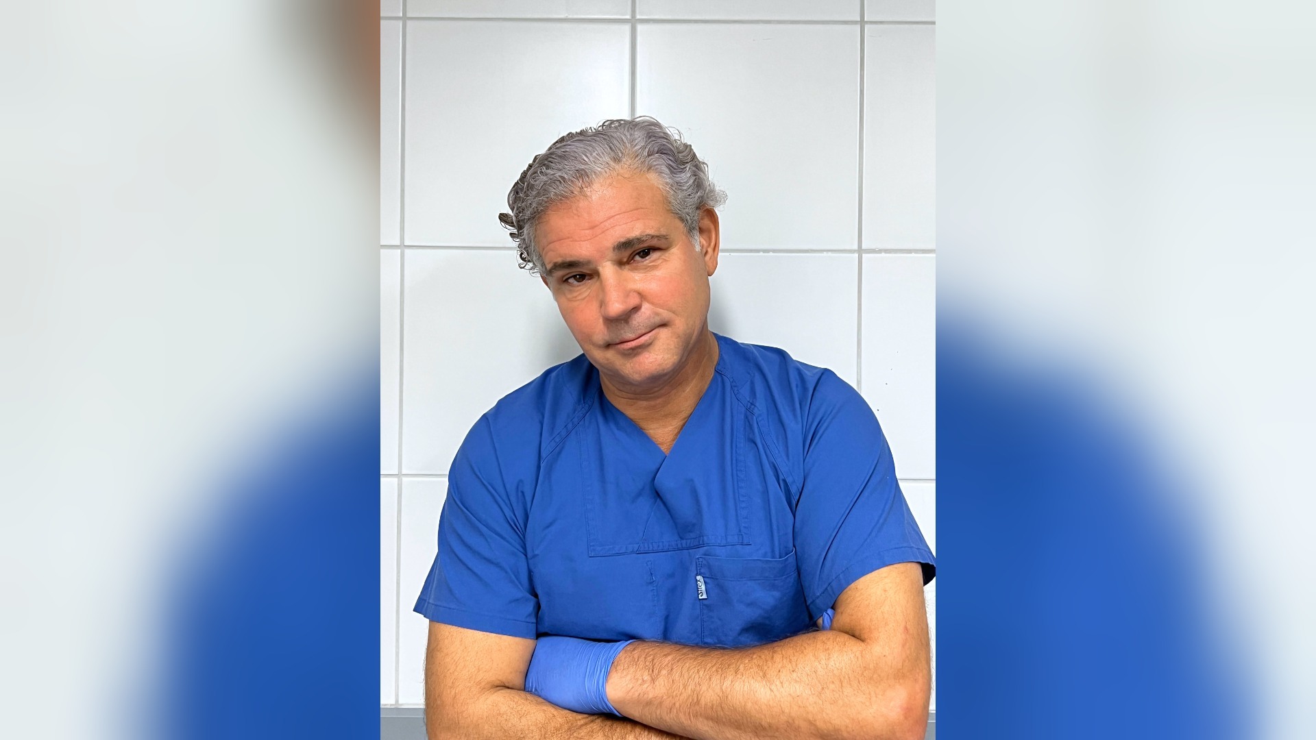 Dr. Michael Tsokos in blauer Arztkleidung und blauem Handschuh mit verschränkten Armen vor einer weißen Fliesenwand