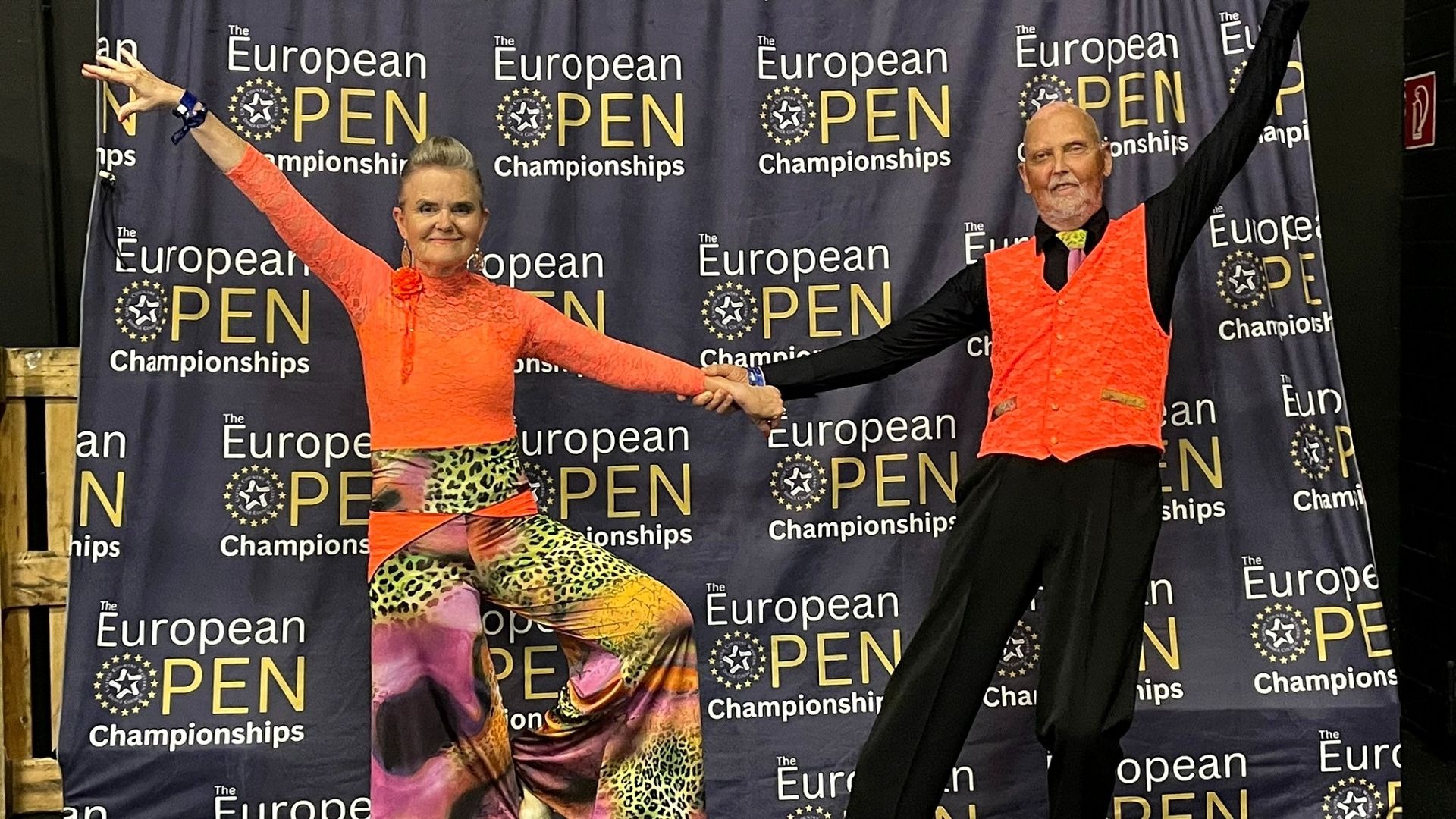 Ehepaar Stüwe in orangefarbenen Tanzoutfits hält Hände, die Arme erhoben, lächelnd vor einer blauen Meisterschafts-Wand
