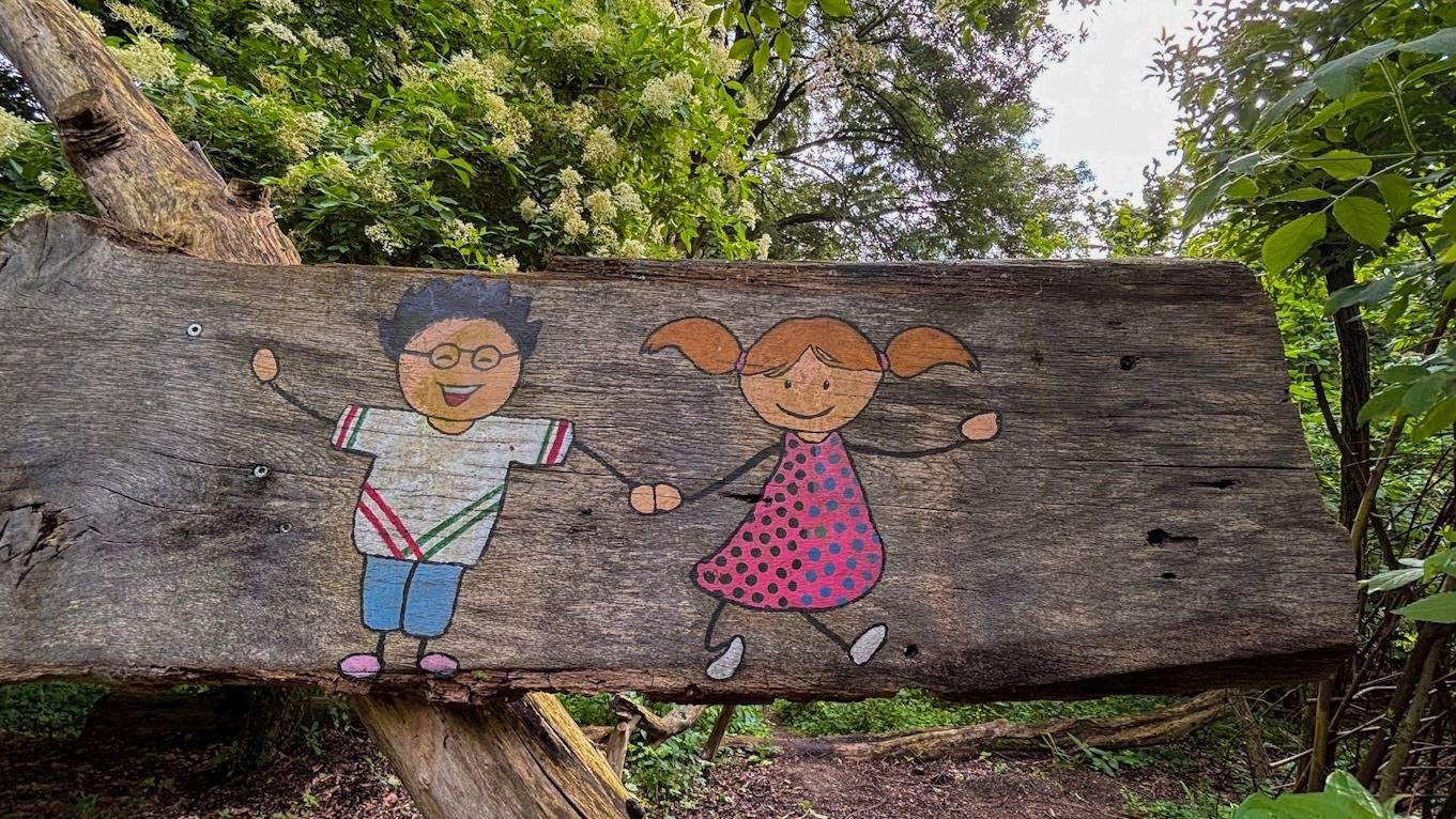 Holzschild mit aufgemalten, lächelnden Kindern, einem Jungen mit Brille und einem Mädchen mit Zöpfen, im Wald